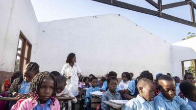 A teacher stands in front of students seated at desks in a partially roofless classroom with white walls, under daylight.