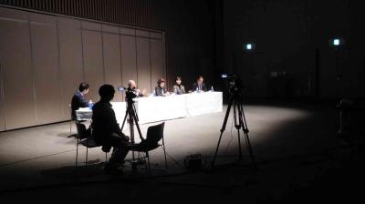 A panel of four people sits at a table with microphones under spotlights, while a camera operator films and another person sits nearby in a dimly lit room.