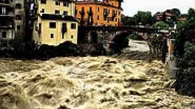 Floodwaters surge through a river in a town, with turbulent water nearly reaching buildings and a bridge in the background.