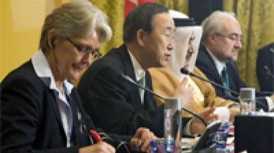 Four people in formal attire sit at a conference table with microphones and papers, participating in a panel discussion or diplomatic meeting.