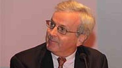 An older man wearing glasses, a suit, and a tie is looking to his left against a plain background.