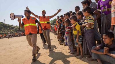 Two aid workers wearing orange vests direct a group of children standing in line on a sandy ground under clear sky. One worker holds a megaphone.