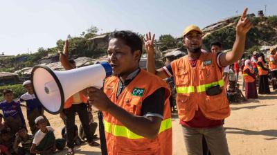 Two men in orange vests use a megaphone and hand signals to address a crowd outdoors, with several people seated and some buildings in the background.