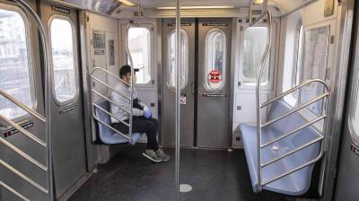 Person wearing a mask and gloves sits alone on a subway train, with empty seats around them.