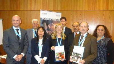 Eight people in business attire stand together indoors, some holding documents, in front of a wooden wall and a banner that reads "WORDS INTO ACTION.