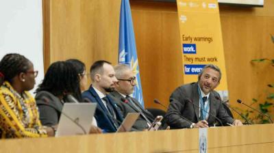 Five people sit at a conference table with microphones, engaging in discussion; a banner behind them reads, "Early warnings work. They must work for everyone.