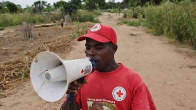 A man wearing a red Red Cross shirt and cap speaks into a megaphone on a dirt path in a rural area.