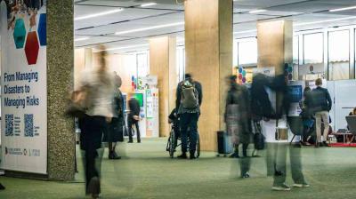 People walk through an indoor event space with informational booths and banners, including one that reads, “From Managing Disasters to Managing Risks.”.