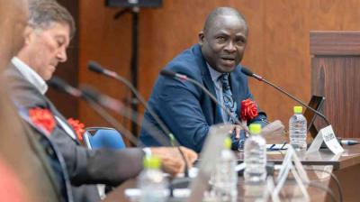 A man in a suit speaks at a conference table with microphones, while another man listens. Bottled water and nameplates are visible on the table.