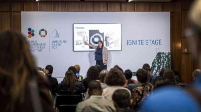 A speaker presents in front of an audience on the Ignite Stage at the Global Platform for Disaster Risk Reduction in Switzerland, with a cartoon slide displayed behind them.