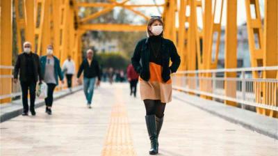 People wearing face masks walk on a yellow-painted pedestrian bridge during the daytime. A woman in front wears a short dress, boots, and a mask. Trees are visible in the background.