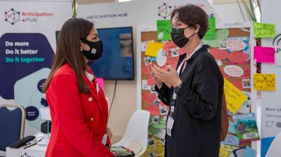 Two women wearing masks and conference badges have a conversation at an exhibition booth with colorful sticky notes on a board behind them.