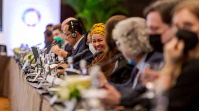 People seated at a long conference table participate in a formal meeting; microphones and headsets are visible, and one woman in a yellow headscarf is smiling.