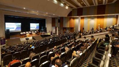 People sit in a large auditorium with tiered seating, facing a stage with screens displaying presentation slides at a conference or seminar.