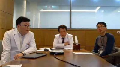 Three men are seated at a conference table in an office; two wear white lab coats, and one wears casual clothes. There are papers and a laptop on the table.