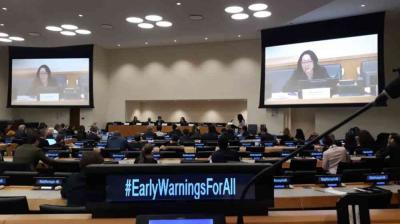 Conference room with attendees seated at desks, a speaker shown on screens, and "#EarlyWarningsForAll" displayed prominently in the foreground.