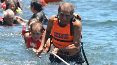 Several people wearing life jackets are pulled through water by a rope during a rescue operation, with one older man leading the group.