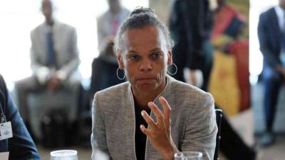 A woman in a gray blazer sits at a table, gesturing with her hand while speaking. Several people are seated in the blurred background.