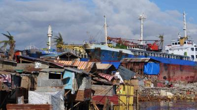 Cargo ships grounded near makeshift homes in a coastal area, with debris and palm trees visible under a partly cloudy sky.