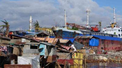 Large cargo ships are docked behind makeshift homes and debris in a coastal area with cloudy skies and scattered palm trees.