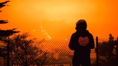 Una figura recortada con un casco y una chaqueta de Santa Cruz se encuentra junto a una valla, frente a un cielo naranja intenso al atardecer.