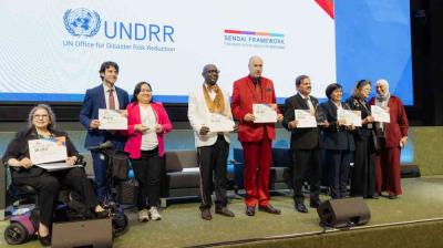 A group of eight people pose on stage holding certificates at a UNDRR event, with the UNDRR and Sendai Framework logos displayed on a screen behind them.