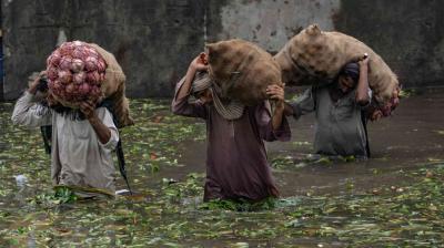 Three people wade through knee-deep floodwater carrying large sacks of vegetables on their shoulders.