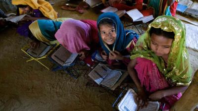 Children in colorful clothing sit on the floor reading from books, with one child looking up and smiling.