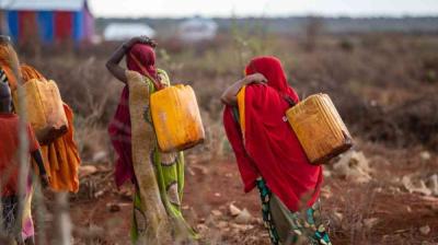 Two women walk through a dry, open area carrying large yellow water containers on their backs, wearing colorful clothing and head coverings.