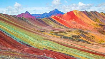Multicolored striped hills and mountains under a partly cloudy sky, showcasing the natural hues of Rainbow Mountain in Peru.