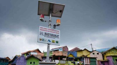 A solar-powered early warning system for floods stands above colorful houses under a cloudy sky.
