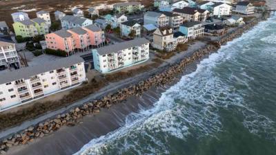 Aerial view of beachfront apartments and houses with waves crashing against a rocky shoreline in a coastal town.