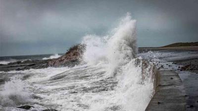 Large ocean waves crash against a rocky shoreline and concrete seawall under a cloudy sky.