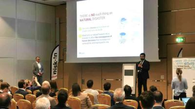 A man speaks at a podium in front of an audience, with a presentation slide titled "There is no such thing as natural disaster" displayed on a screen behind him.