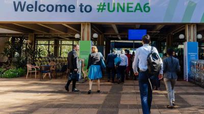 People walk toward an entrance under a “Welcome to #UNEA6” sign at what appears to be a conference or event venue.