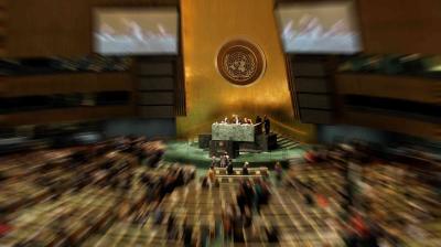 A large assembly room with people seated and a central podium, featuring the United Nations logo on a gold backdrop.