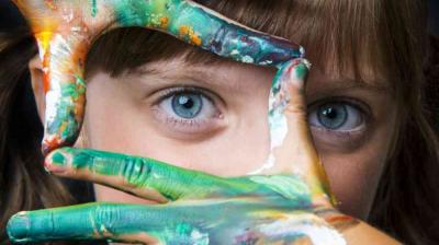 Child with blue eyes frames face with hands covered in multicolored paint, focusing on the eyes.