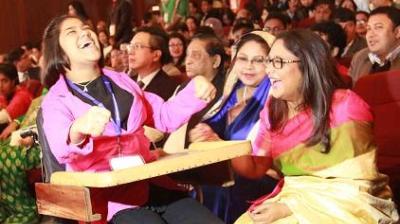 Two women are laughing joyfully in a crowded auditorium. One woman is wearing a pink jacket and seated in a wheelchair with a tray. The audience is engaged and smiling.