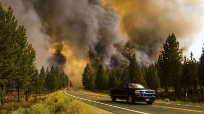 A dark smoke cloud from a wildfire looms over a forested area next to a road, with a black pickup truck in the foreground.