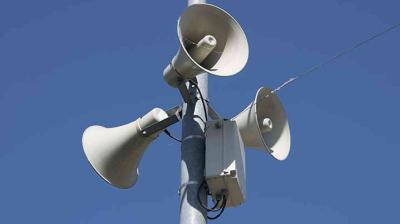 Three loudspeakers attached to a metal pole against a clear blue sky.