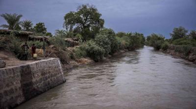 A wide canal with muddy water flows past dense vegetation and simple structures; two people stand on the bank next to yellow jerry cans.