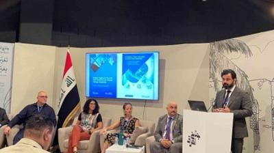 A panel of five people, including one speaker at a podium, is discussing at an event. There is an Iraqi flag and a presentation screen in the background.