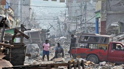 People stand and sit among debris on a street filled with rubble and damaged buildings, with a red vehicle nearby, in the aftermath of a disaster.