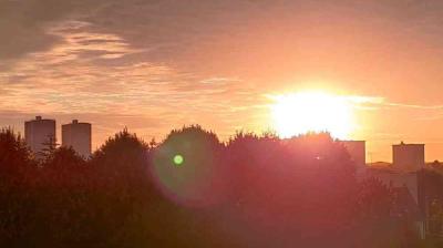 The sun is setting behind trees and buildings, casting a warm orange glow across the sky with a lens flare visible in the foreground.