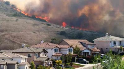 A wildfire burns on a hillside near a residential neighborhood, with smoke and flames visible behind homes.