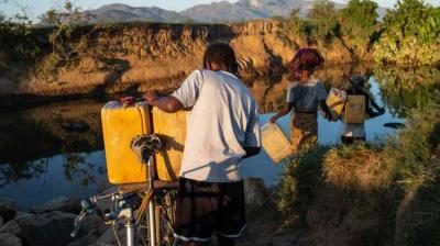 People crossing a river, carrying yellow containers and a bike. The terrain is rocky, and mountains are visible in the background.