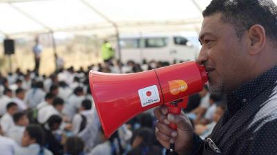 A man speaks into a red megaphone in front of a seated crowd under a large tent.