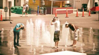 Three people cooling off by leaning over water fountains in a public area on a sunny day.