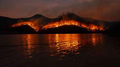 A line of fire burns on a hilly landscape at night, with the flames reflecting on the water in the foreground.