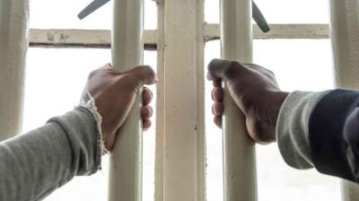 Two hands gripping white metal jail bars, viewed from the inside.